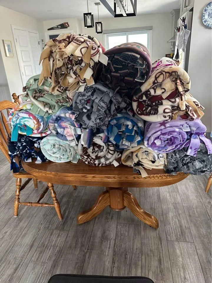 A large pile of finished rolled fleece tie blankets stacked on a dining table, ready to be delivered