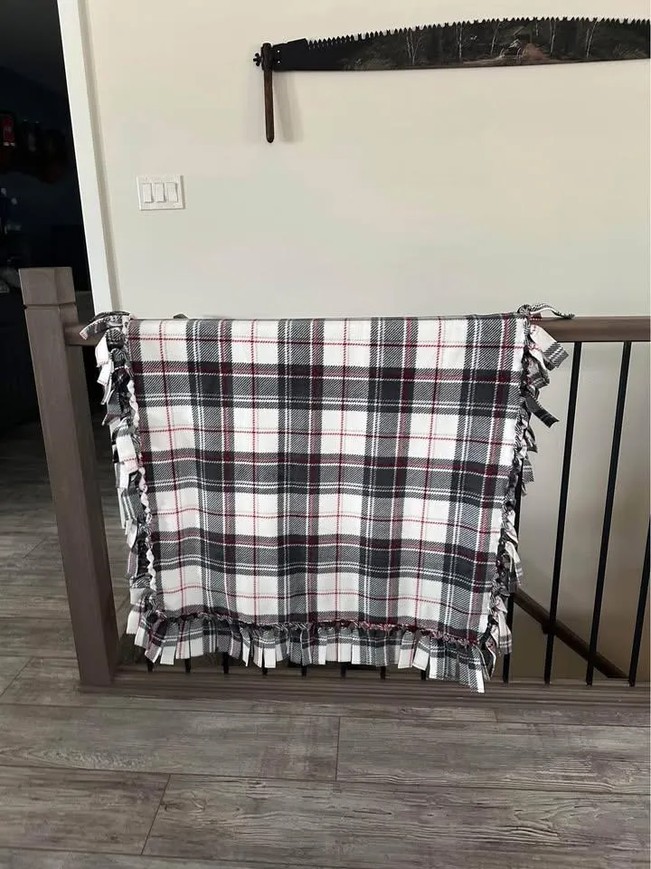 Red, white, and charcoal plaid fleece tie blanket with a knotted fringe hanging over a stair railing
